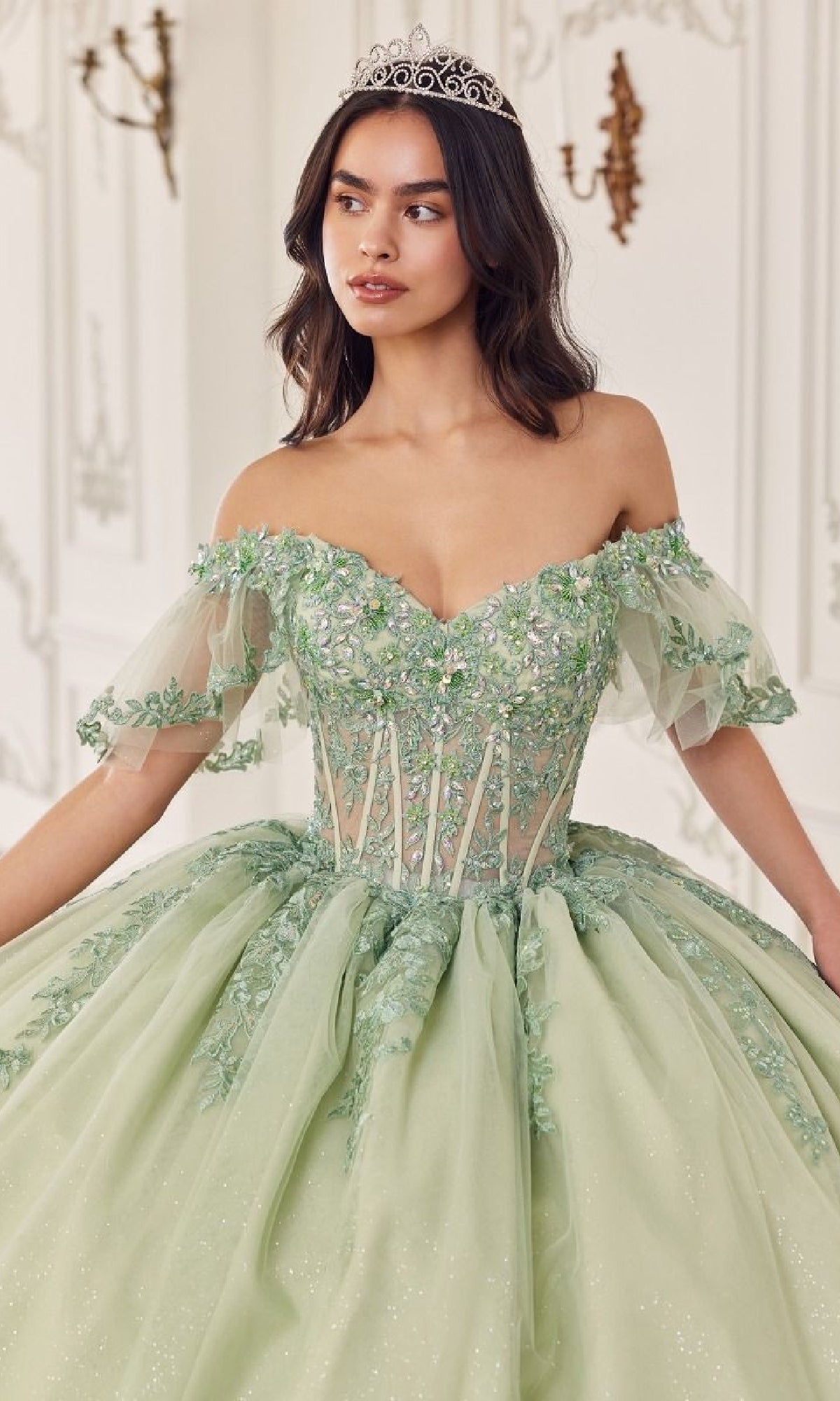 A woman in the Quinceanera Dress 15725 by Ladivine, a sage green off-the-shoulder gown with floral lace appliques and a tiara, stands gracefully in an ornate room, adding elegance to the regal setting.